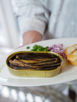 Sardines in a tin with greens and bread on a white plate, perfect for gourmet cuisine.
